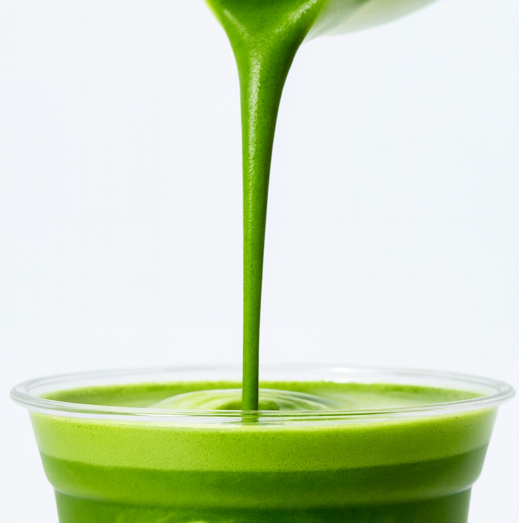 Green smoothie being poured into a clear plastic cup with a white background